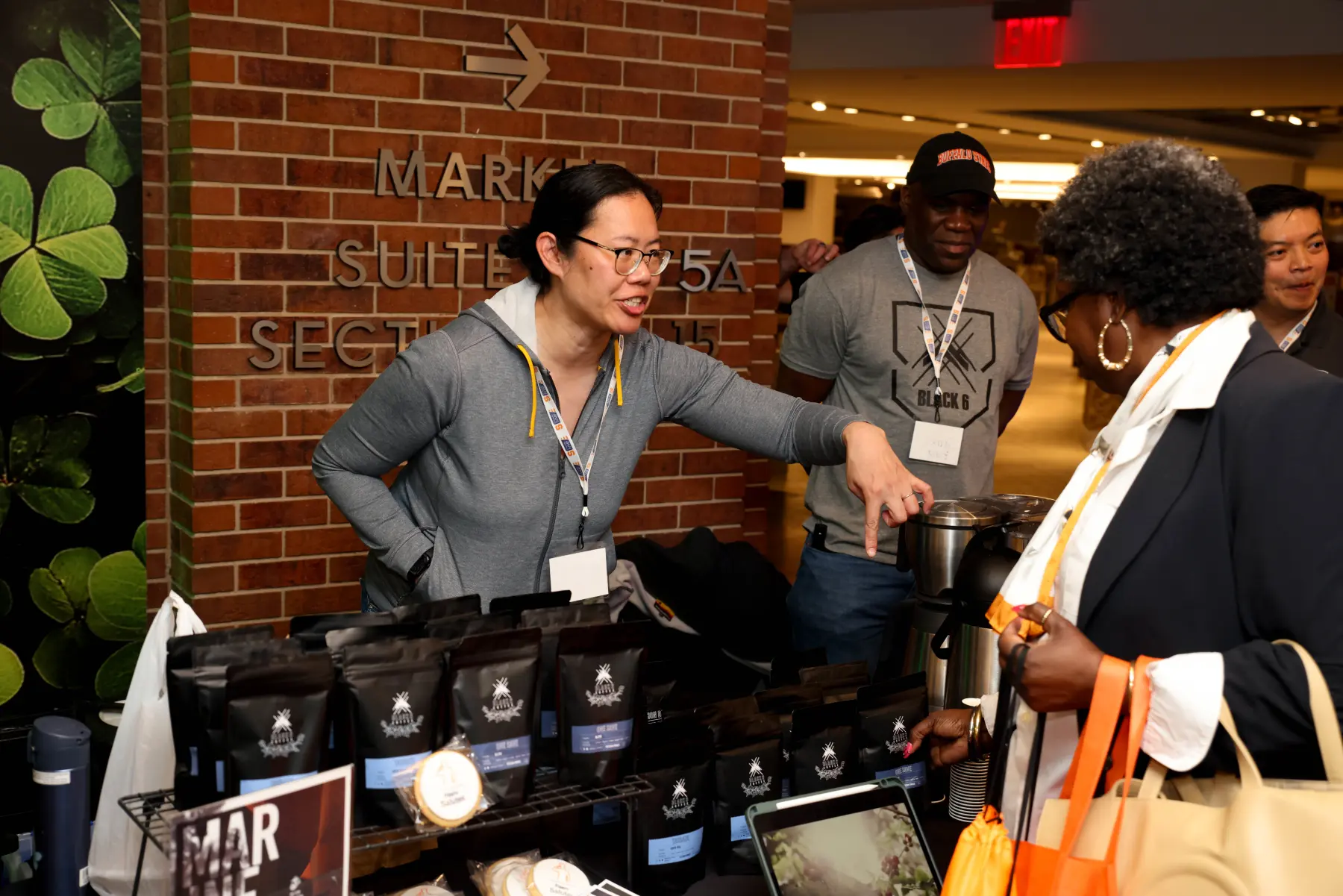 A coffee vendor greets MEF NYC attendees from behind her table, which holds a variety of bagged coffee and large thermos urns for brewed samples.