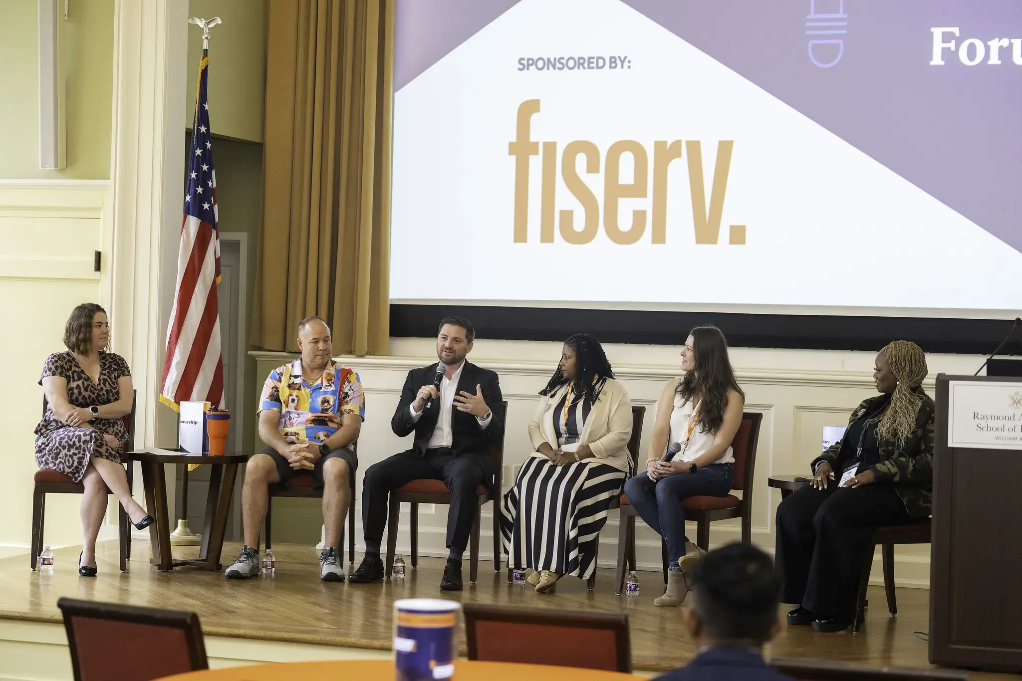 2 men and 3 women sit in a row on stage to the right of the MEF panel session host. One man is speaking into the mic and addressing the audience.