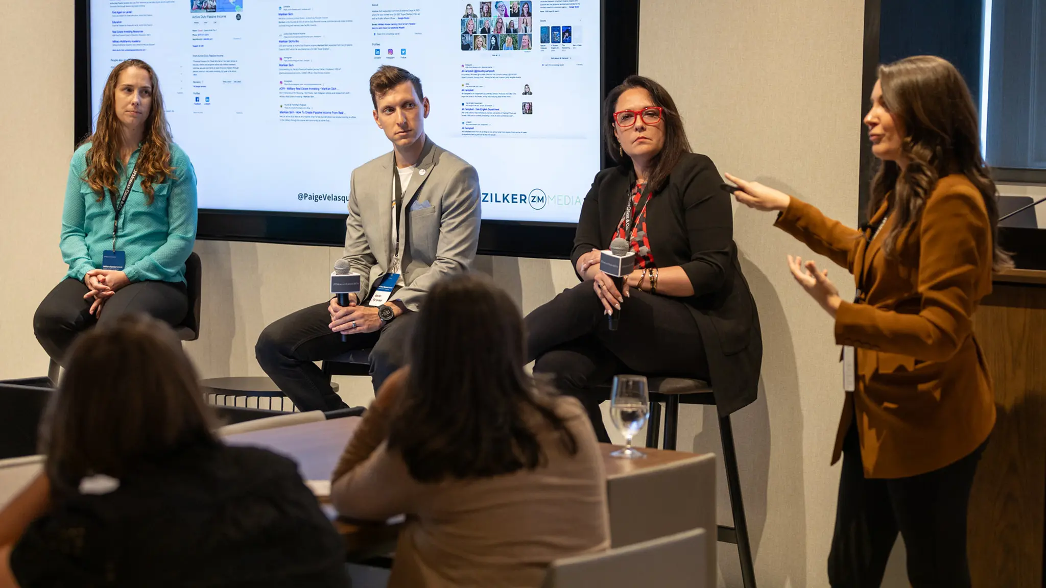 A group of 3 panelists sit on tall stools in front of a crowded room while the session host addresses the MilSpouse Summit 2023 crowd.
