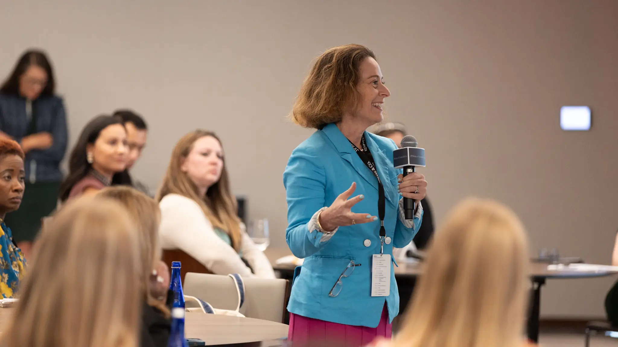 A woman wearing a blazer holds a mic while speaking to a person out of frame. The other attendees are focused on the suited woman.