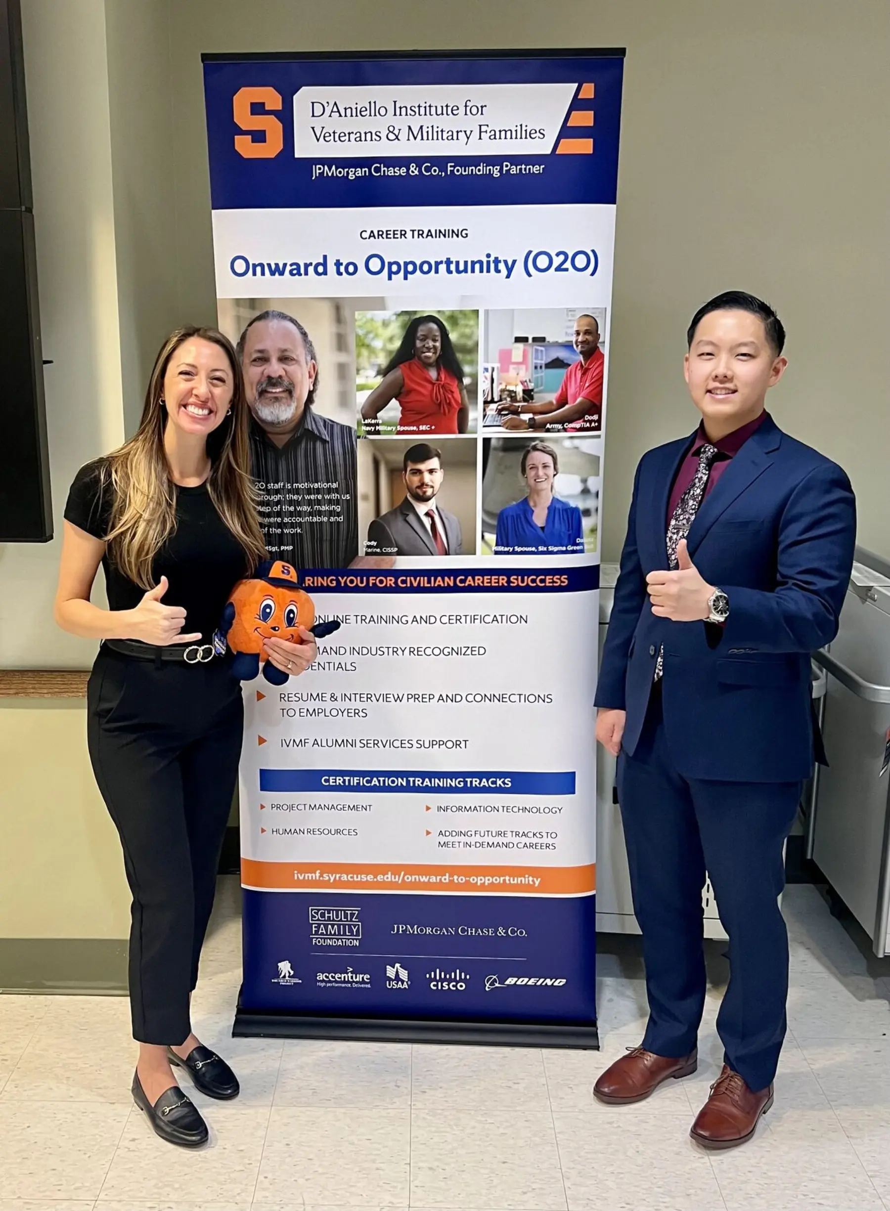 A woman and a man in business attire stand alongside a Syracuse IVMF O2O stand-up banner giving thumbs-up. The woman is holding a plush Orangeman.