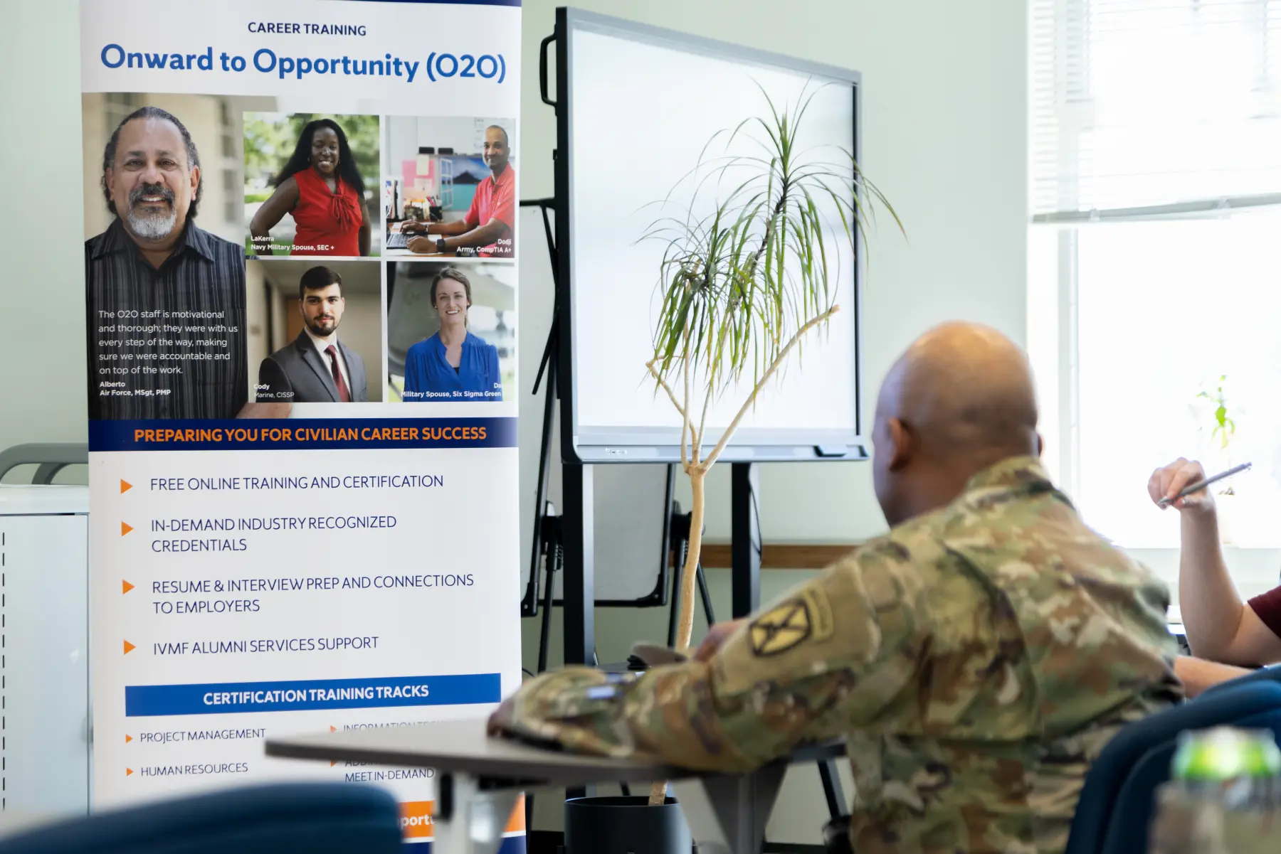 A service member in uniform sits at the front of the brightly-lit O2O classroom session with the O2O stand-up banner directly in front of him.