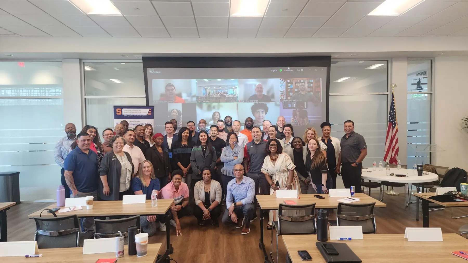 A group of O2O attendees poses together at the front of the classroom with a projection screen including remote attendees.