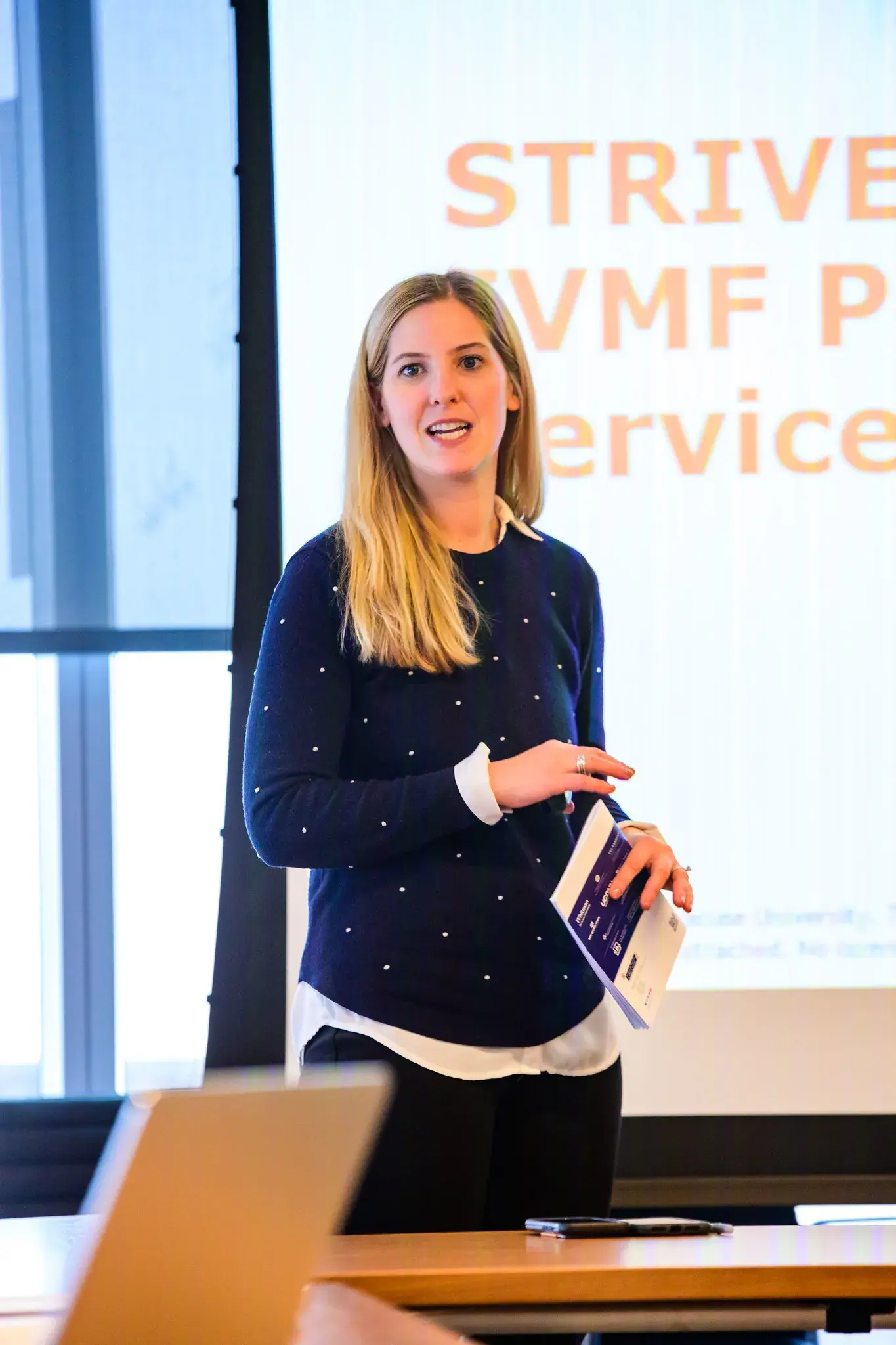 A woman stands between a table and presentation screen at a STRIVE event in a large conference room.