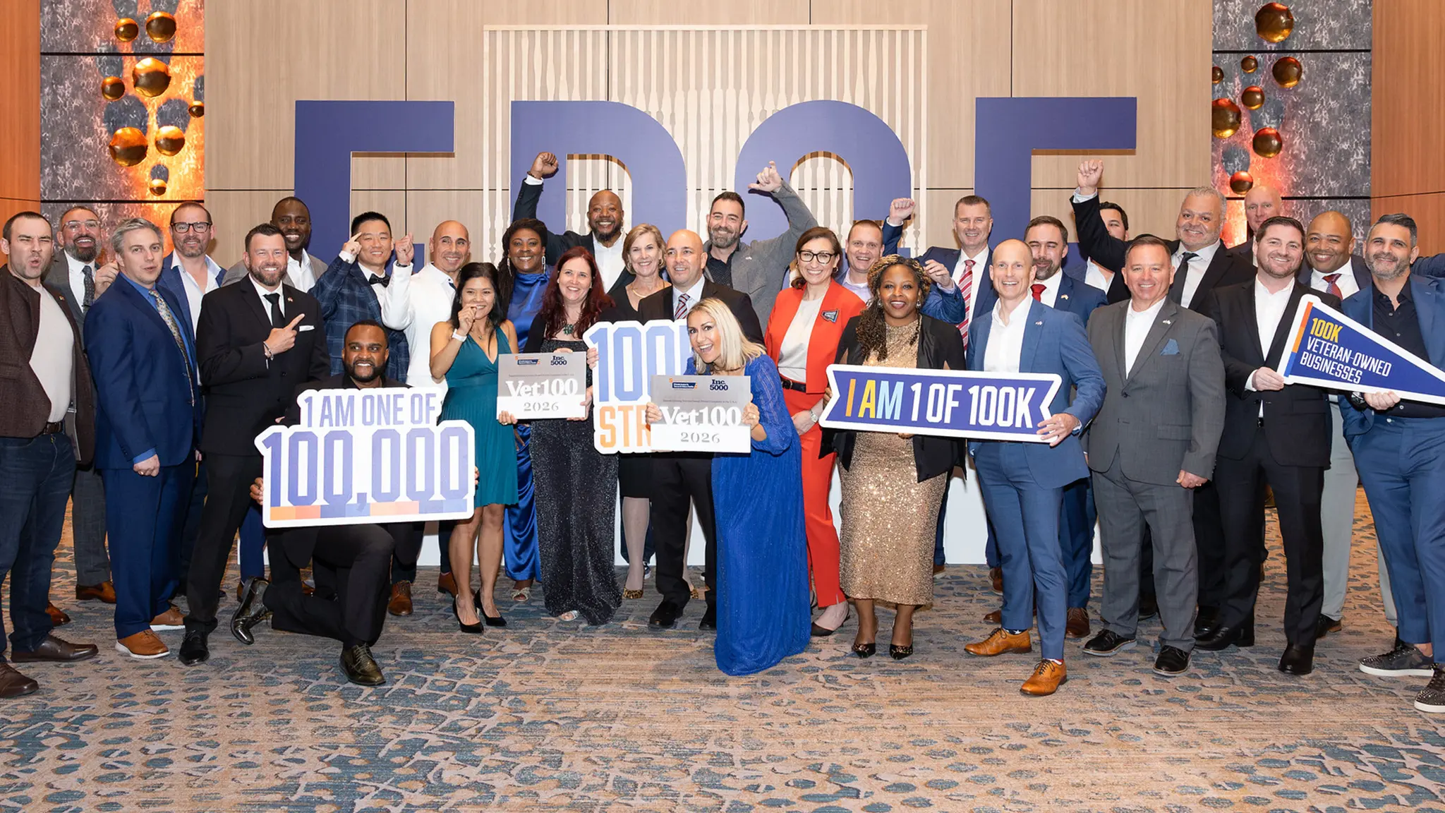 A group of 30 Veteran Edge 2026 attendees stands together smiling in front of the large E D G E letters. Some are holding signs for Vet100 and "I am 1 of 100K".