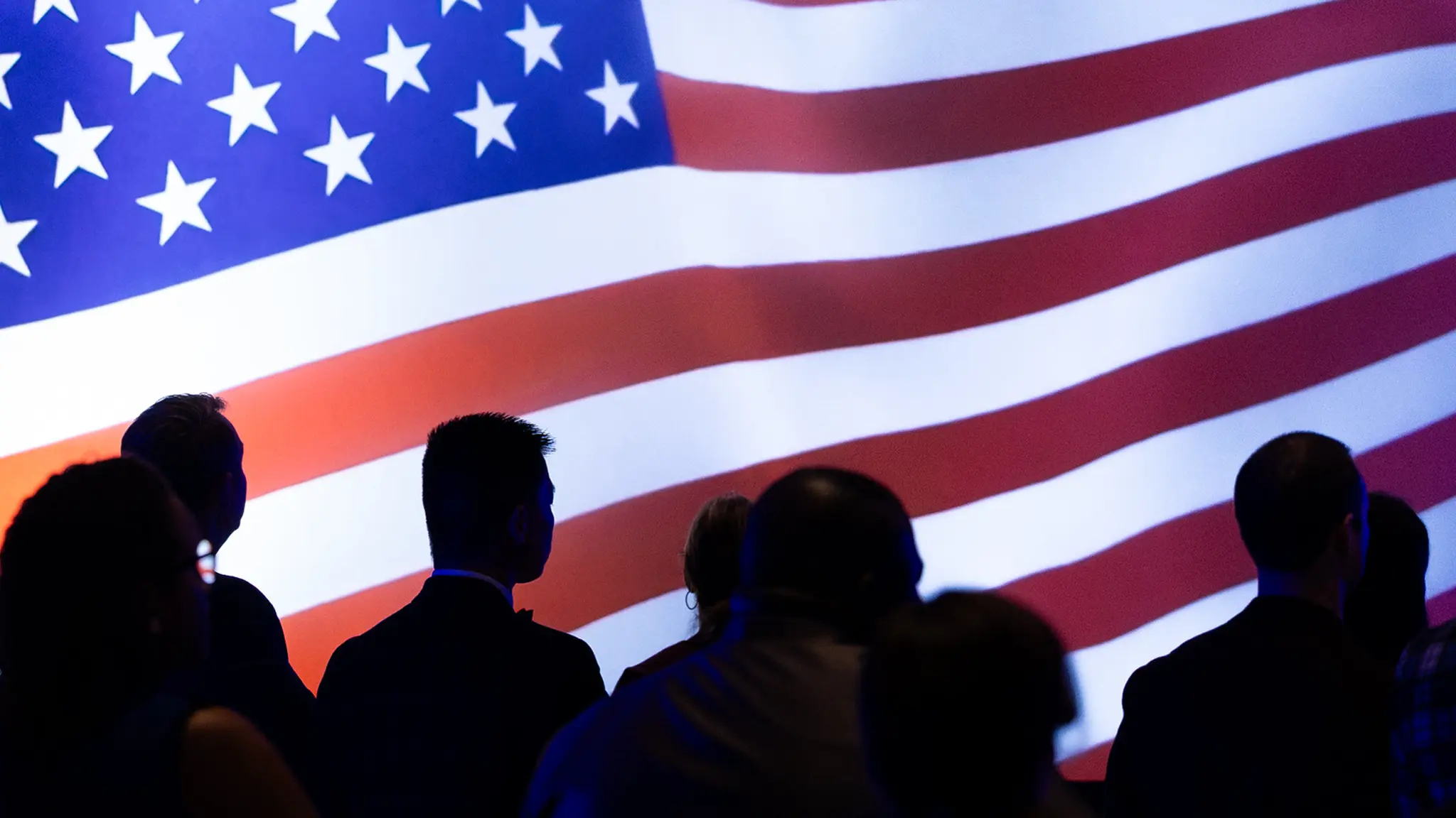 Silhouettes of the upper portion of people from behind. All are in front of a large bright display screen featuring a gently waving American flag.