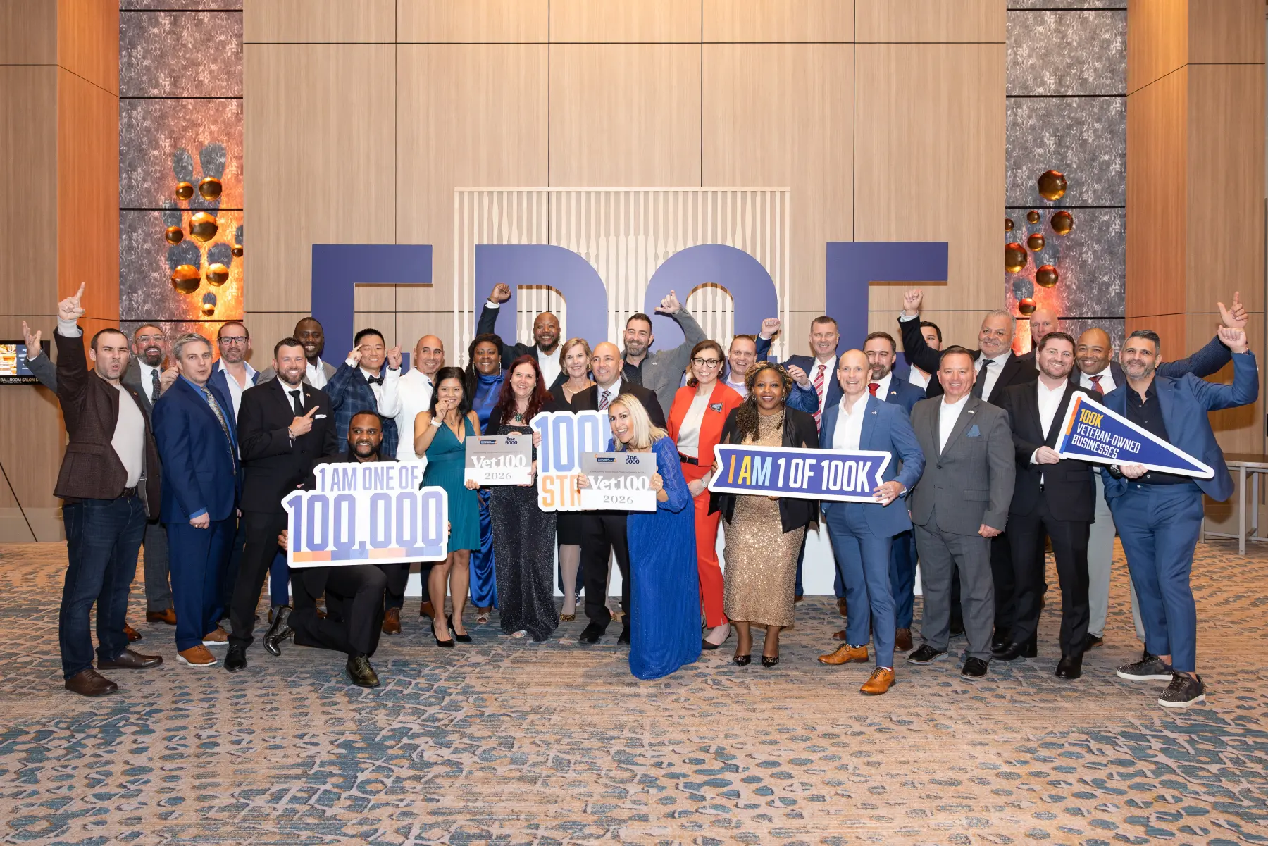 An excited group of Veterans Edge 2026 attendees pose together in front of the large E D G E letters and hosing 100K signs.