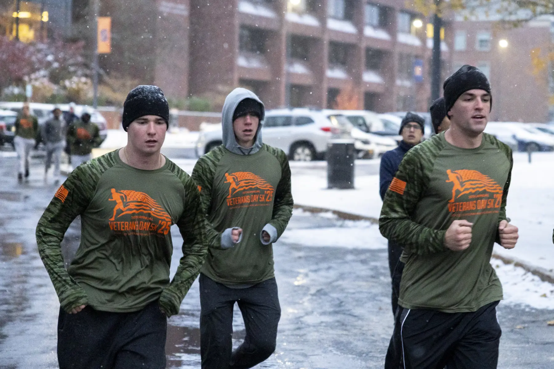A group of 3 runners pass by the camera, mid-stride, sporting camo patterned Veterans Day 5K long-sleeve shirts and frosted with snow.