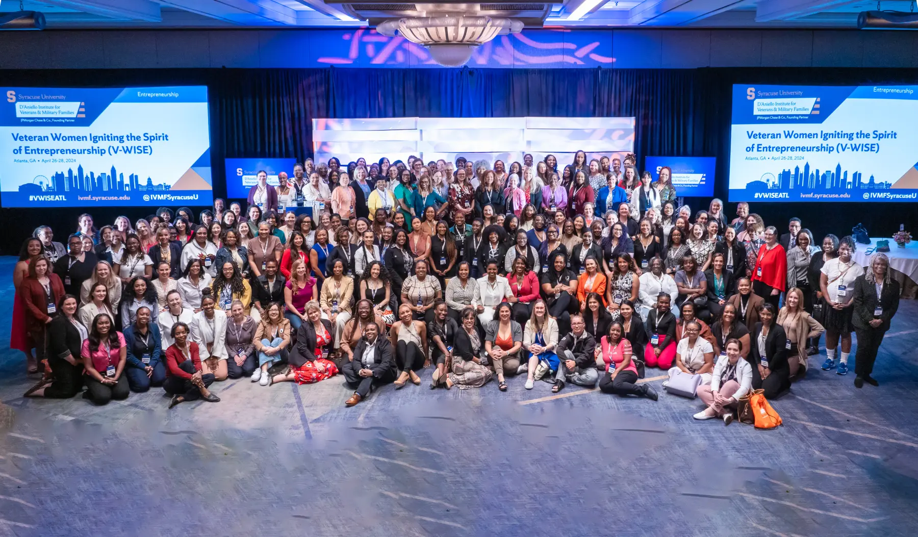 Large group of all attendees a posing together with smiles in front of a the stage in the main event room of a V-WISE event.