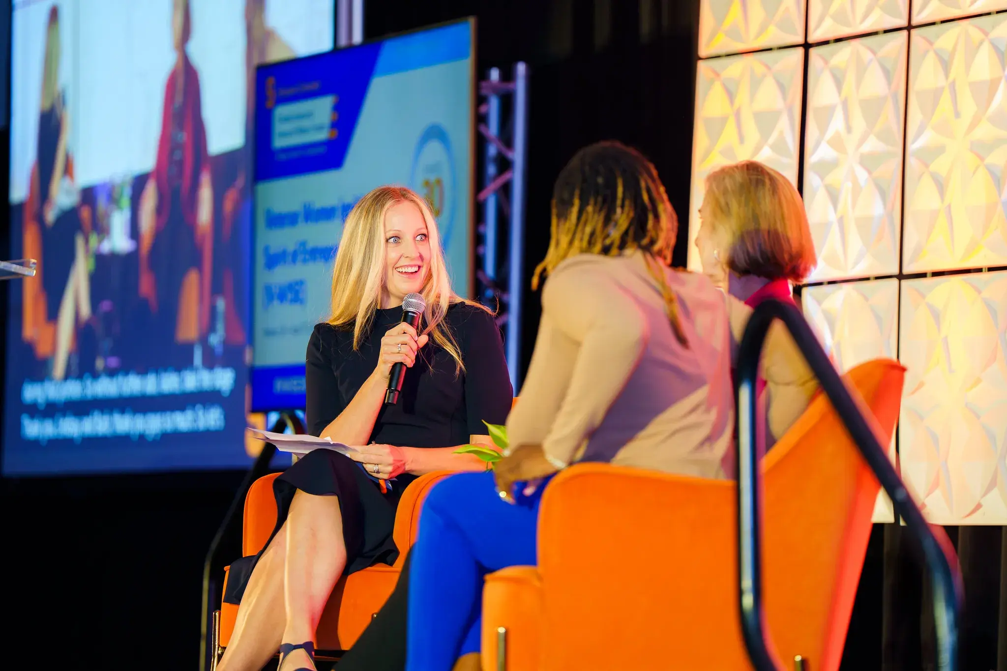 At V-WISE Atlanta, a woman serving as host of a fireside chat on stage looks wide-eyed and smiling at one of the other two on-stage women.