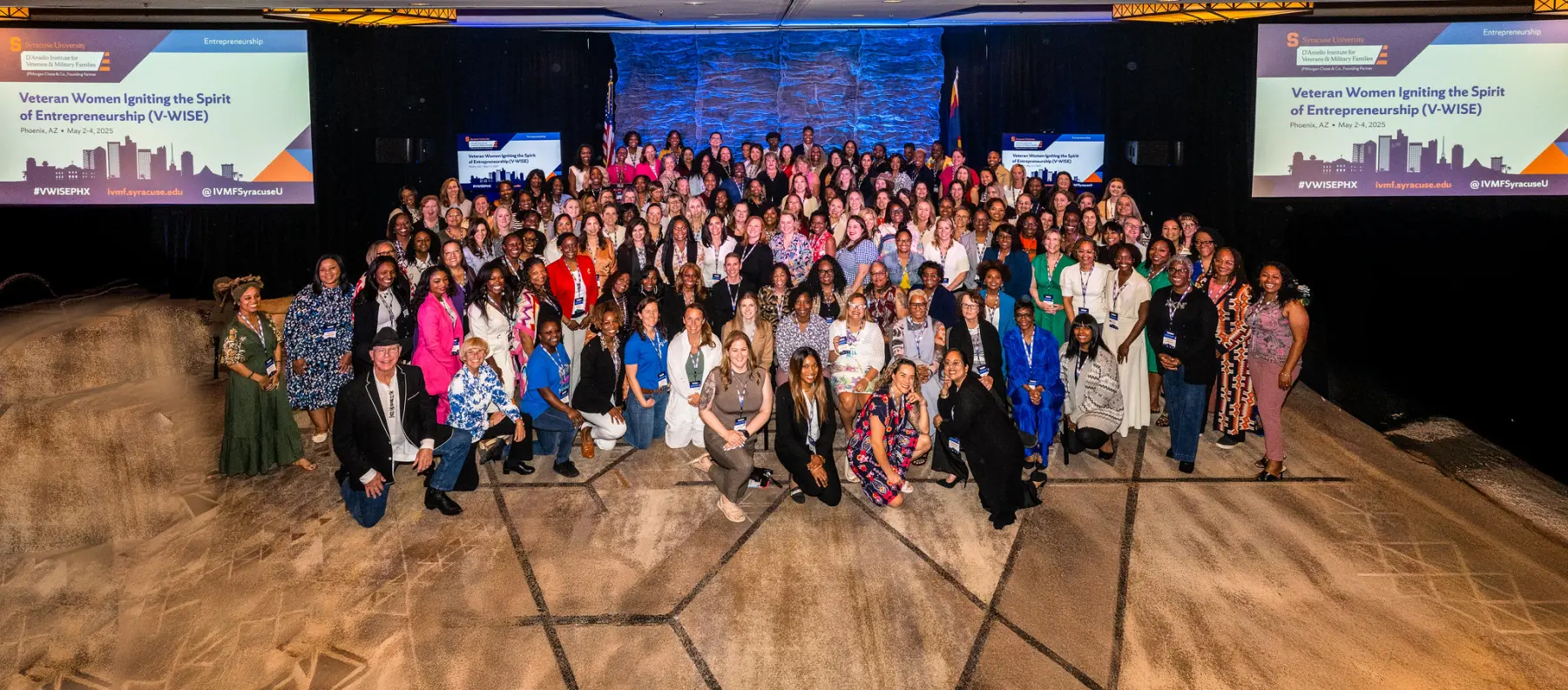 Large group of all attendees a posing together with smiles in front of a the stage in the main event room of a V-WISE event.