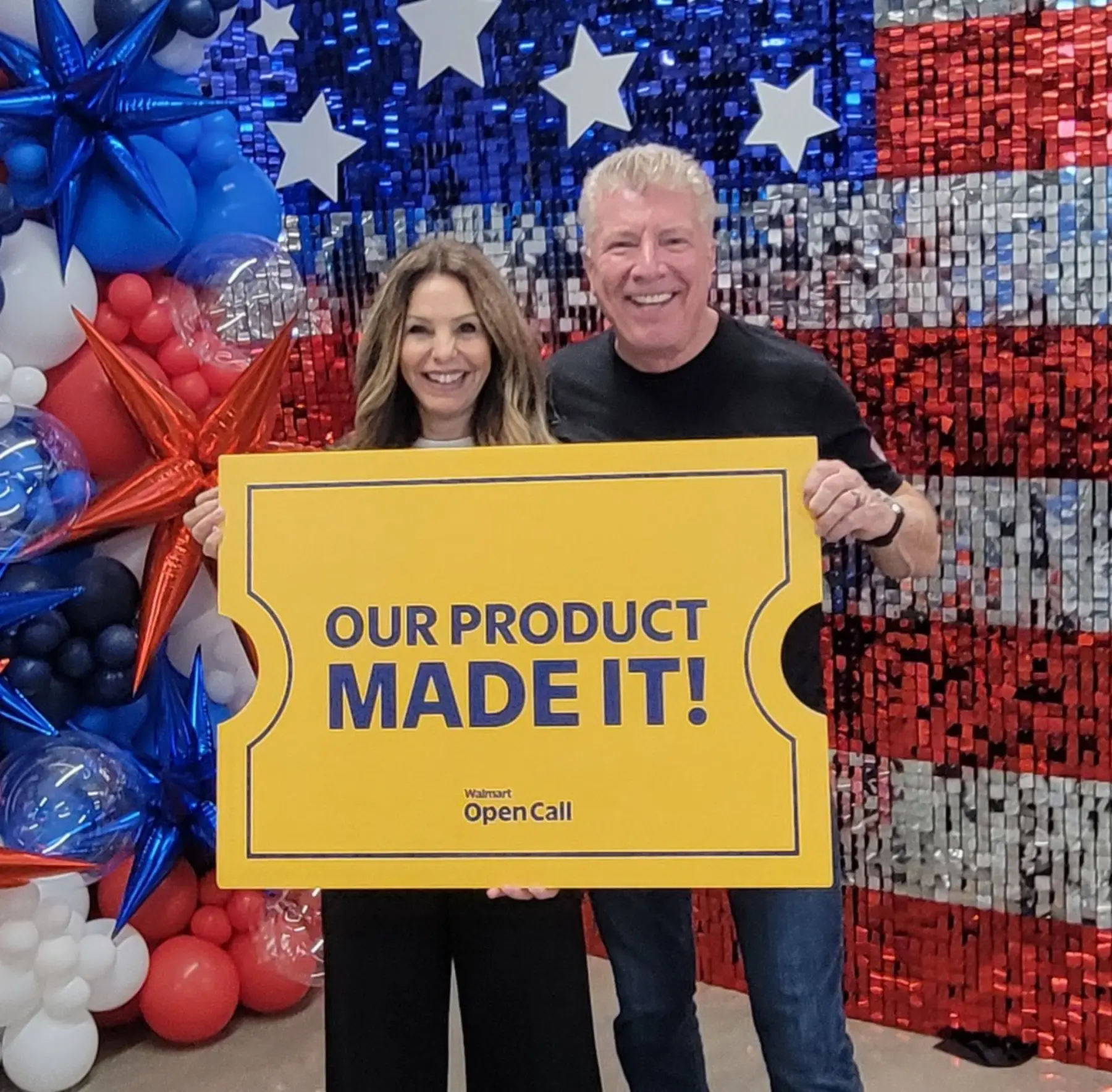 A man and a woman stand together in front of a shimmering American flag backdrop holding a large ticket-shaped prop that says "Our product made it!"