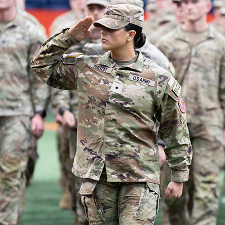 A woman soldier wearing fatigues salutes while marching ahead of a group of soldiers.