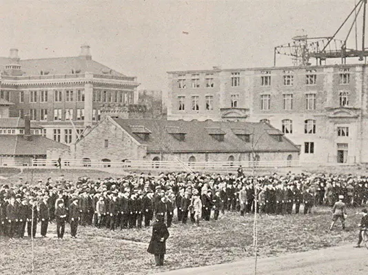 Aged vintage photo showing 4 large groups of troops in formation facing the chancellor in a large open space surrounded by SU buildings..