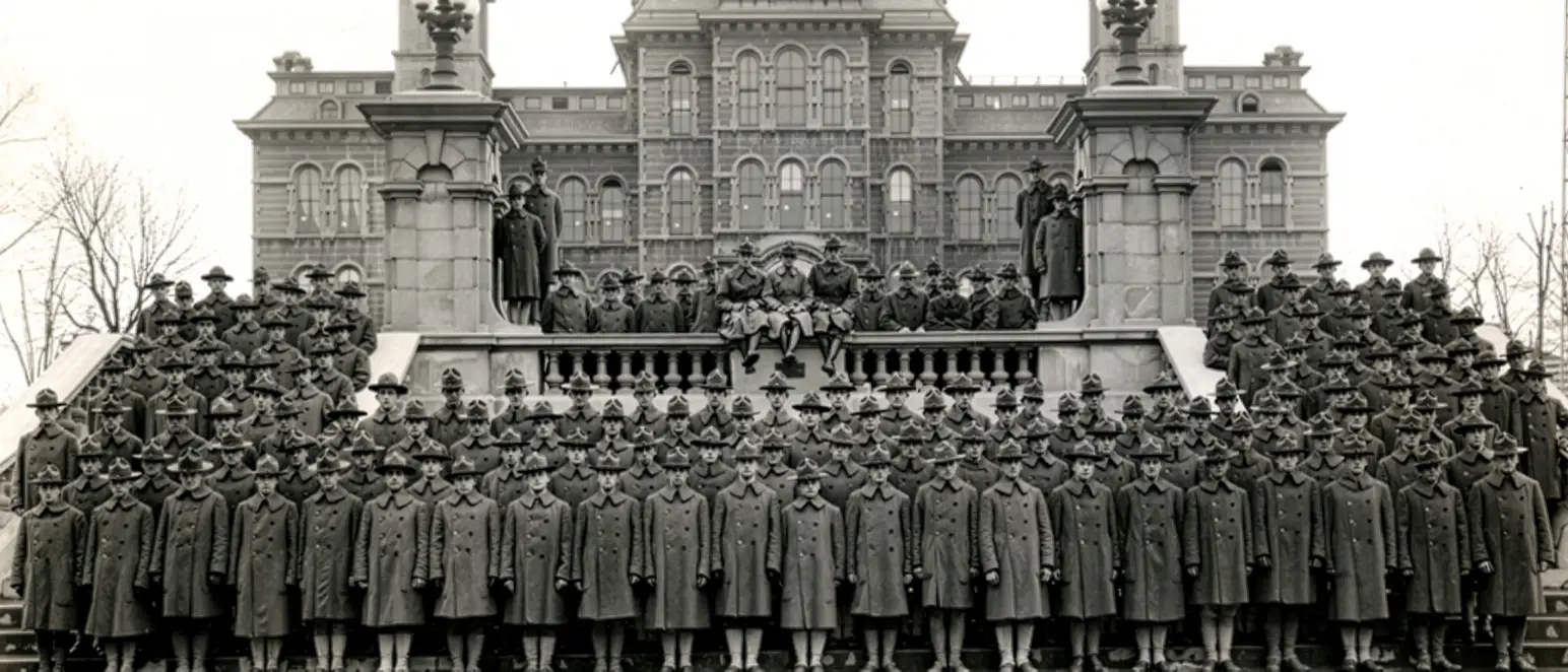 A vintage photo showing a large group of men and women in old uniforms of 21 EVB SU team members stand on the stairway in front of the SU clock tower.
