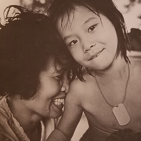 Asian woman presses her face against the arm of a child wearing military dog tag.