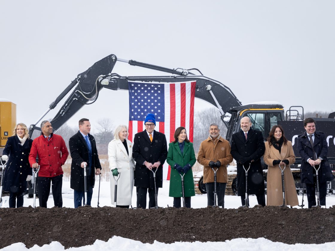 Outdoors on a cold day, 10 people holding shovels stand in a line in front of a crane from which an American flag hangs.