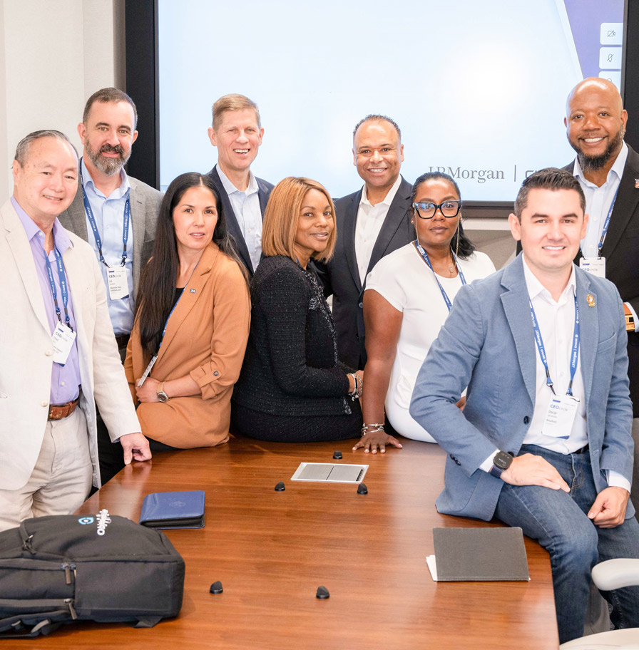 A diverse group of nine smiling CEOcircle participants pose around a conference table in a bright office.