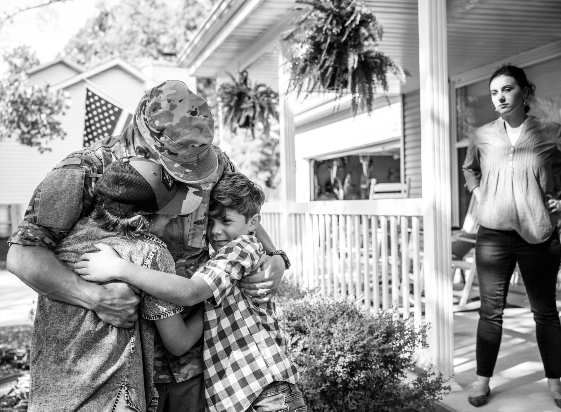 Military spouse on porch watching children hug their father in military uniform