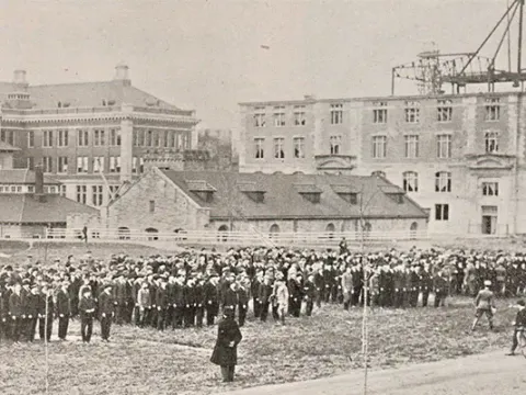 Aged vintage photo showing 4 large groups of troops in formation facing the chancellor in a large open space surrounded by SU buildings..