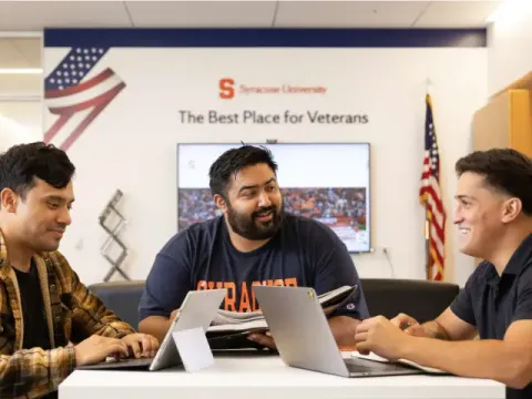 3 men in a SU IVMF room sit around a square table talking while using tablets and computers.