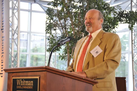 Ted Lachowicz in a suit and tie speaking at a podium with a plaque that reads "Whitman" during the EBV SU 2010 closing ceremony. 