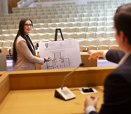 Student Allison Carlos points to an exhibit in a courtroom while wearing a tan blazer, and a man in front of her points to the exhibit