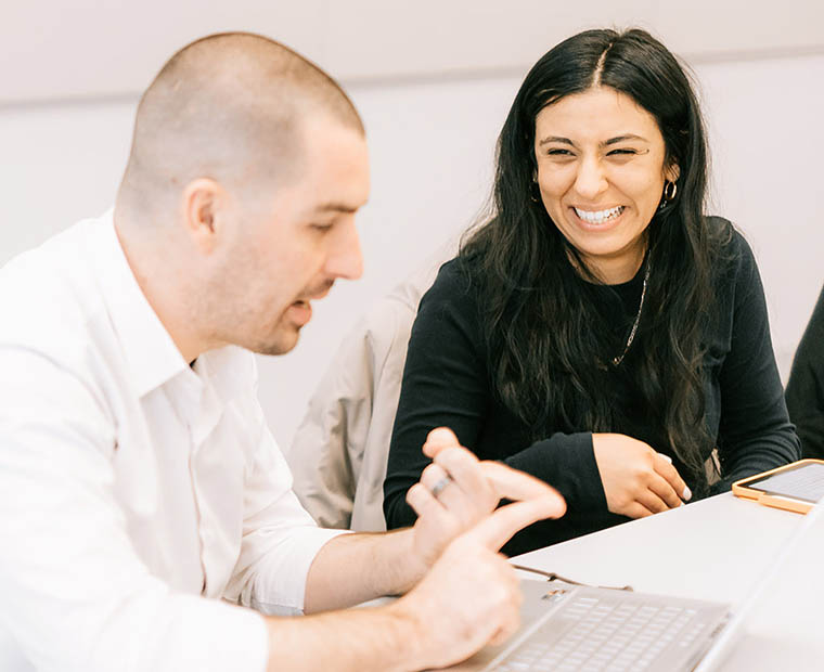 two students work together at a table, smiling in front of their laptops with one looking down and the other looking at him