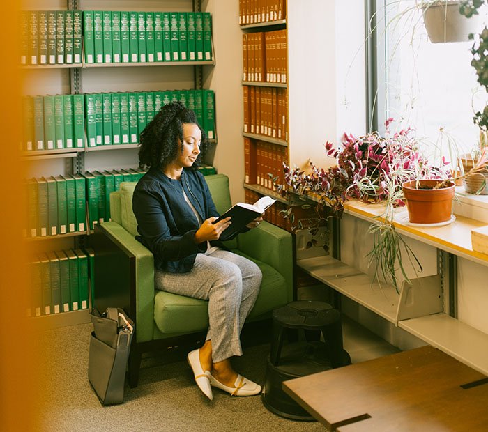 Woman sits in a chair in the law library reading a book, in front of green and brown books on four shelves by the window.