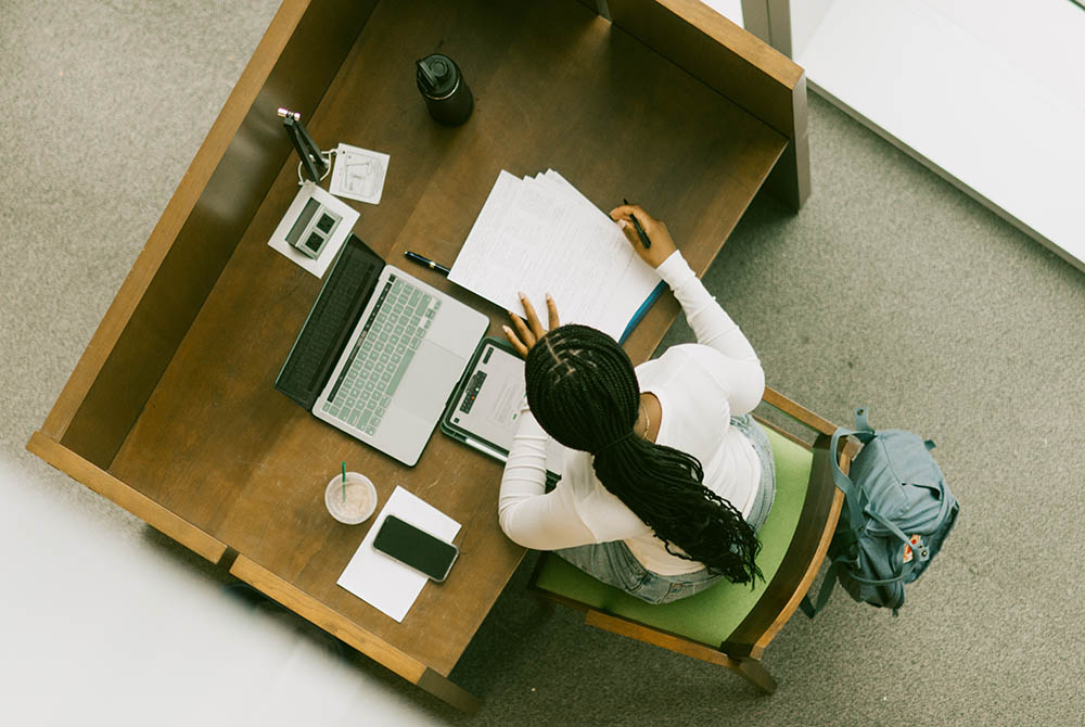 aerial image of a student studying in a library desk, with her laptop in front of her and pages of notes.
