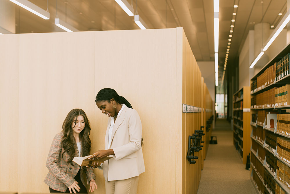 two students look at a book together in the law library, in front of a tan bookcase.