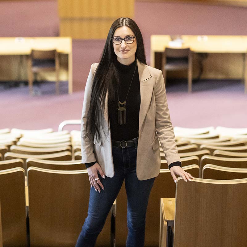 Allison Carlos stands in the empty courtroom with her back to the seats wearing a tan blazer and a black turtleneck