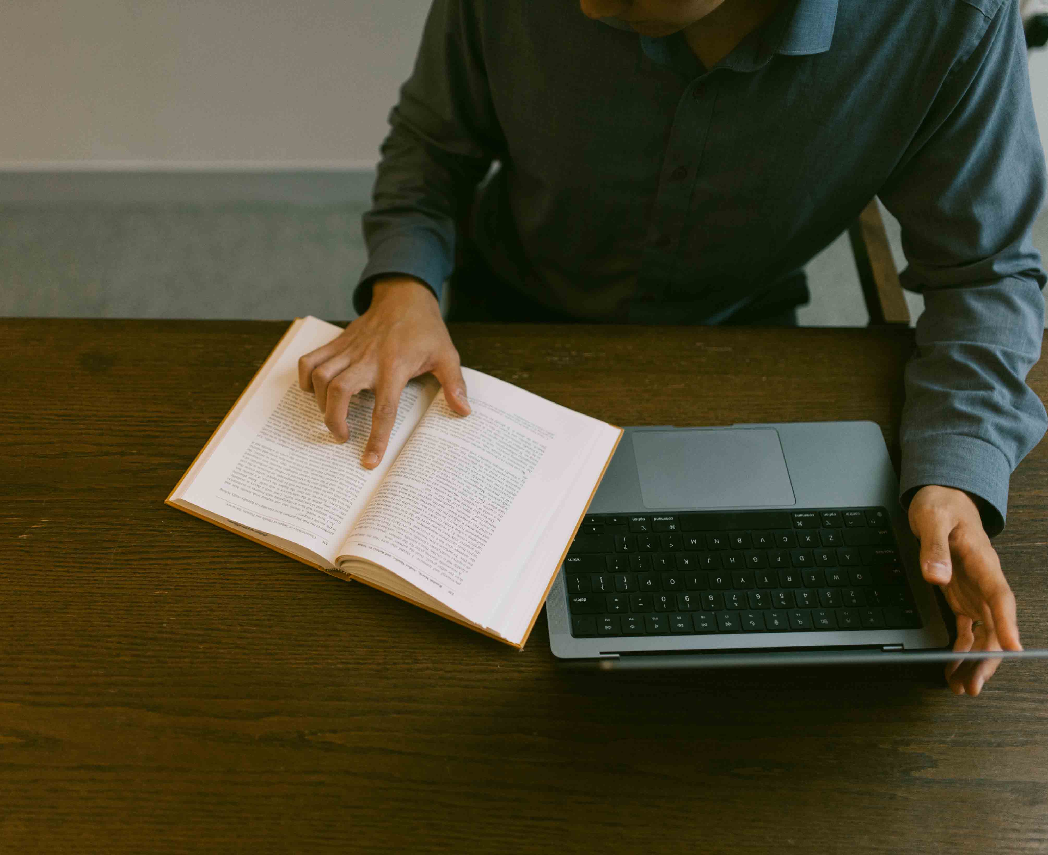 Close up image of a person pointing to a book and a laptop on a desk.