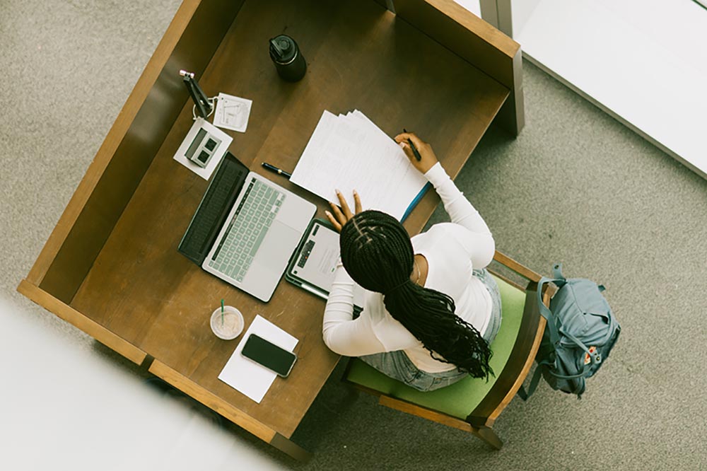 aerial of a student working in the library showing the top of her head, with books and her laptop on the desk in front of her.