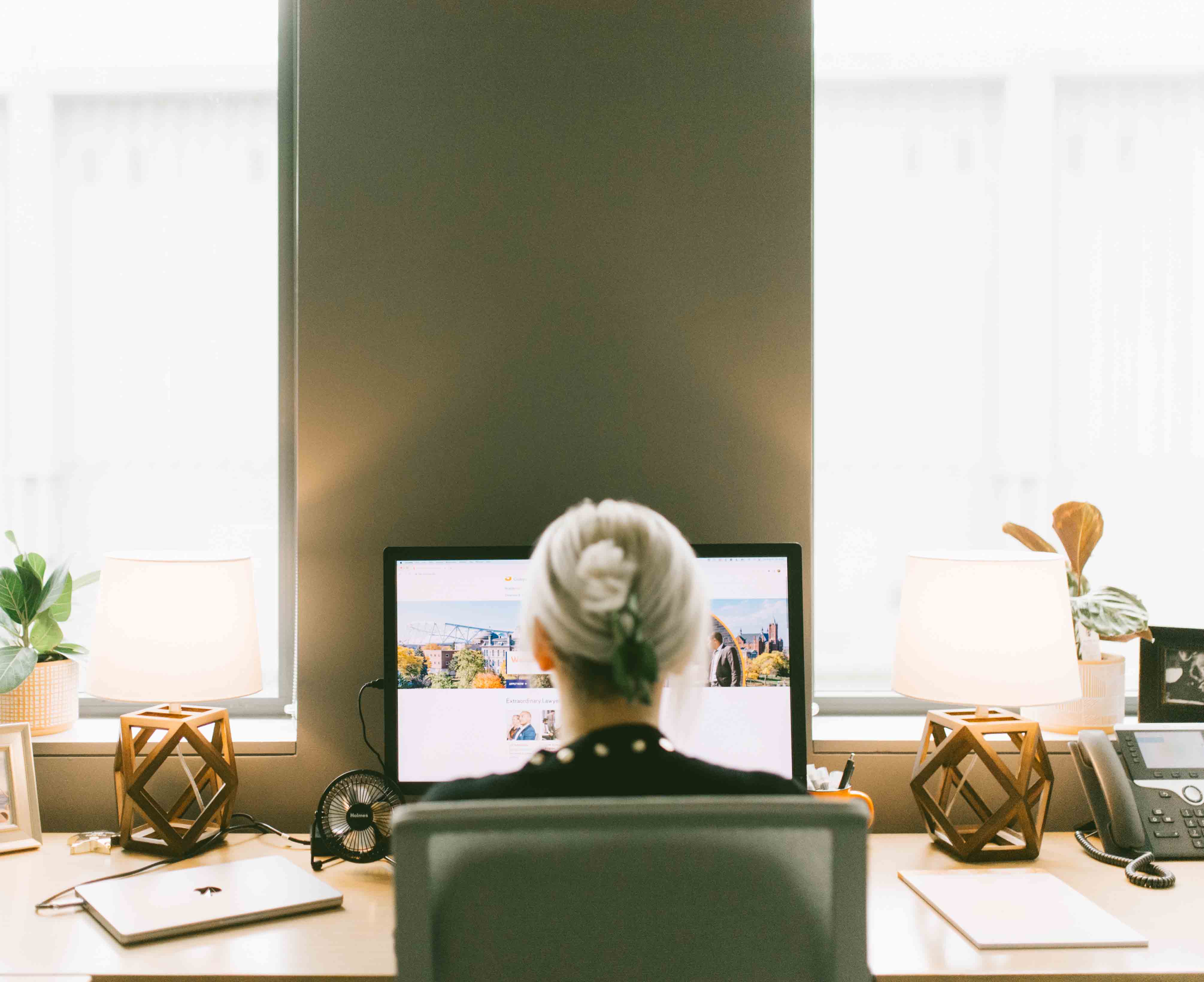 Woman sits at a desk looking at a screen with windows behind the laptop.
