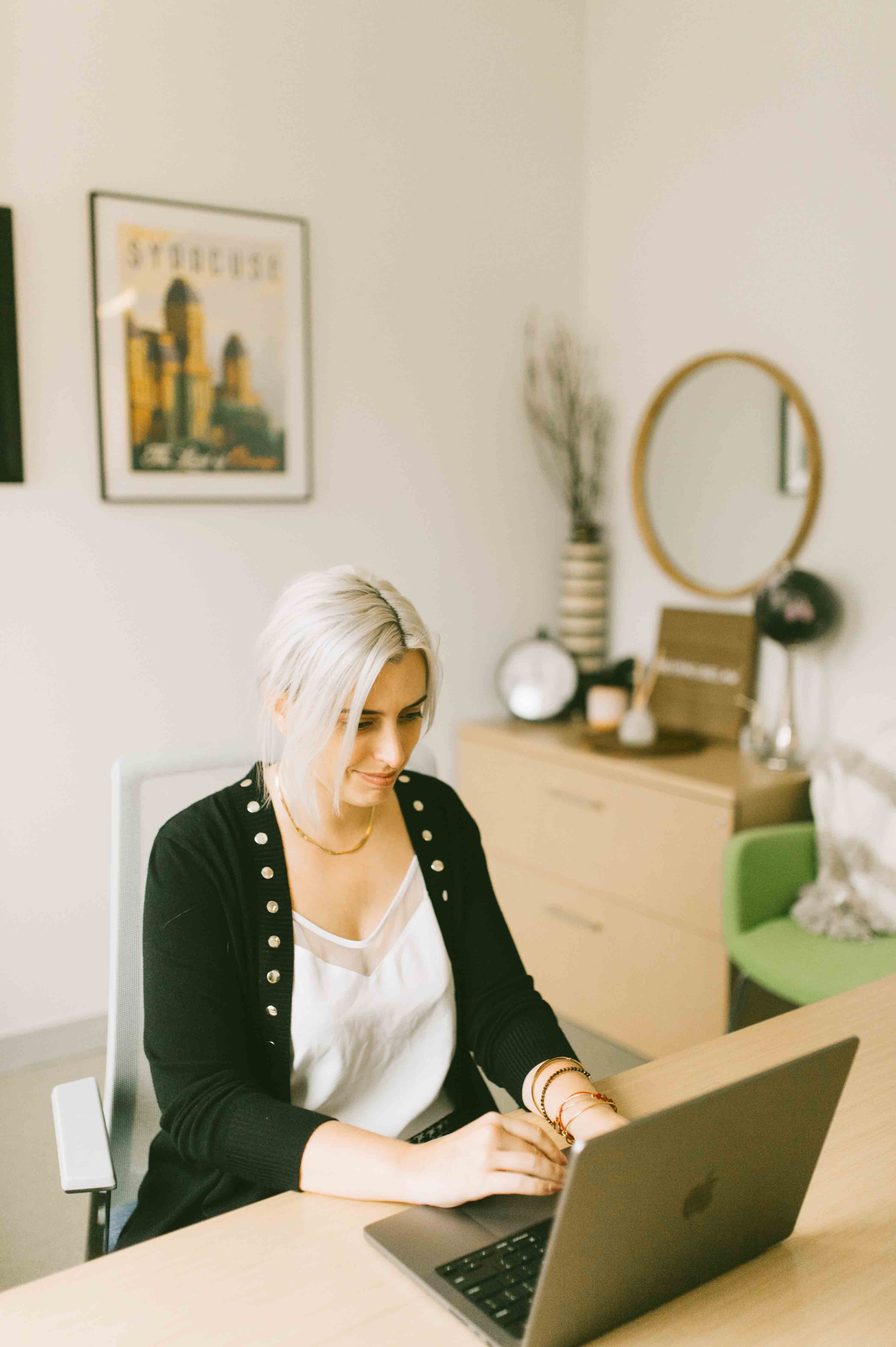 Student works on a laptop in an office, with a syracuse framed photo of the city behind her, wearing a black cardigan over a white shirt.