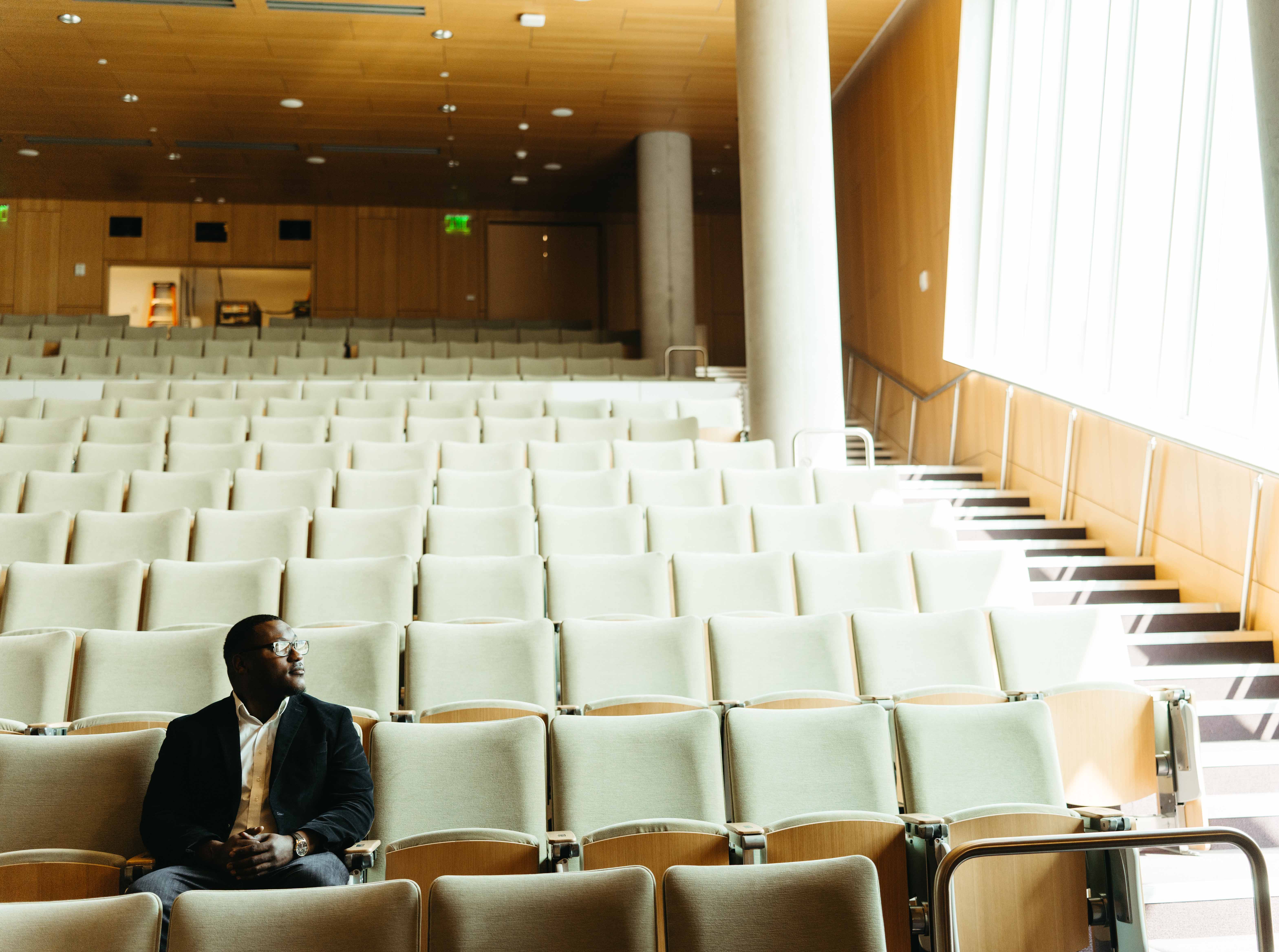 Student sits in an empty courtroom looking out the window, wearing a black suit. 