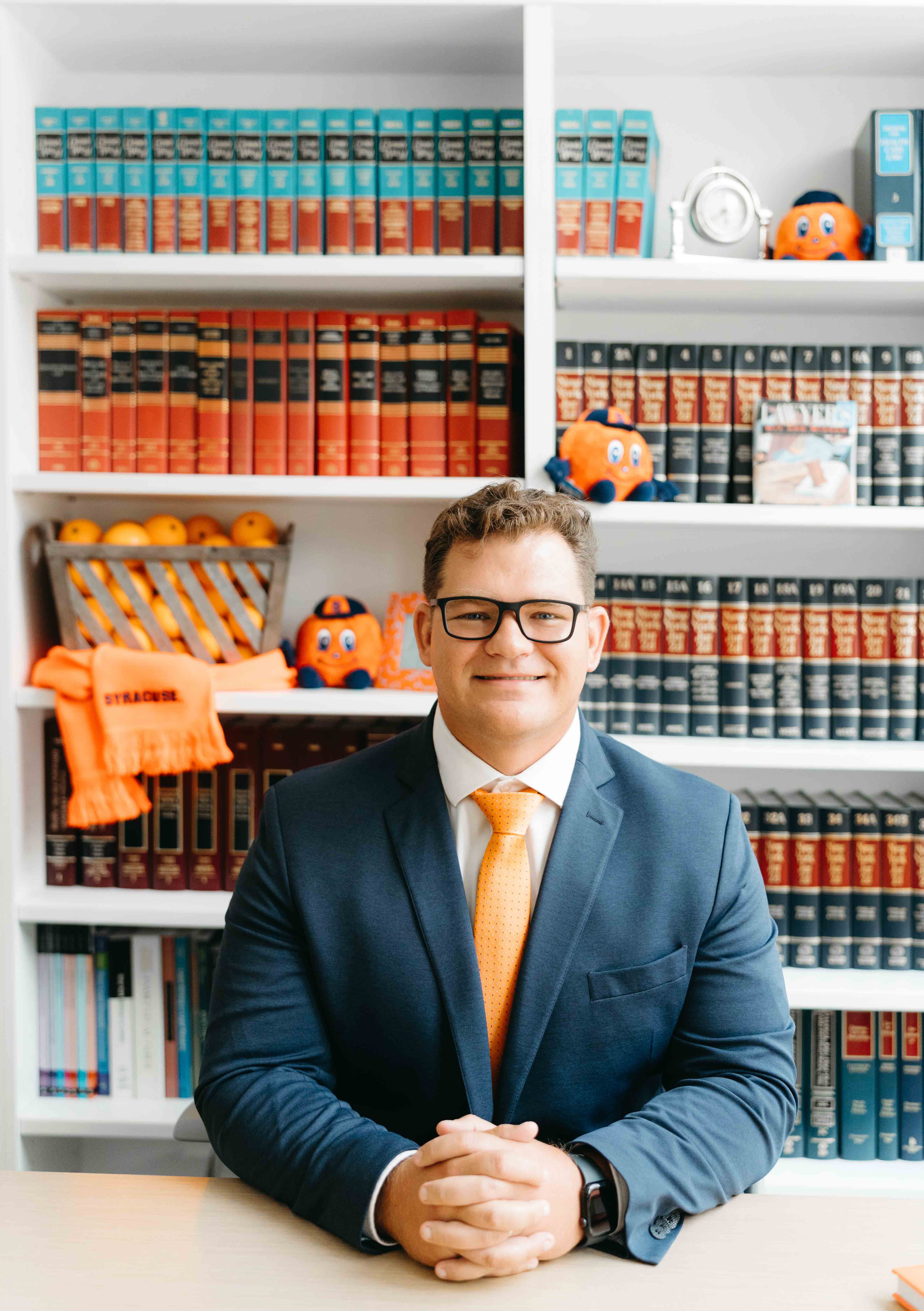 Student sits with a blue suit and an orange tie in front of orange and blue law books, and a cart of oranges on a bookshelf.