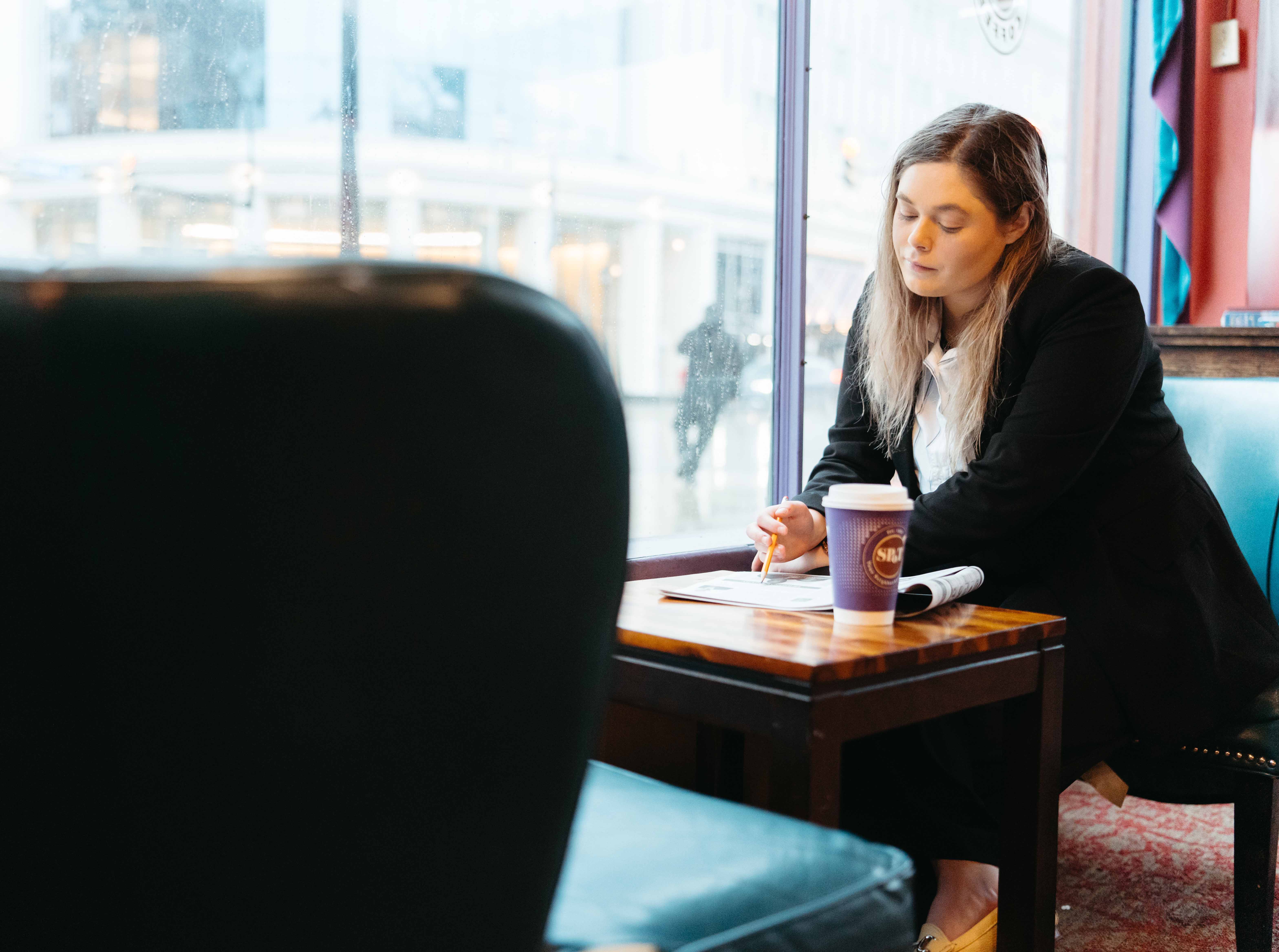 Student sits at a cafe seat with a cup of coffee, looking down at a paper and holding a pen.