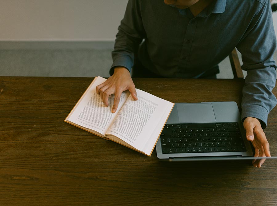 Close up image of a person with one finger on an open book, the other hand holding an open laptop on a wooden desk
