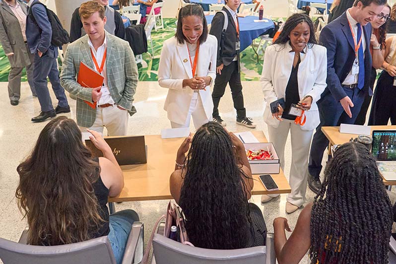 Three students stand in front of a table at a student org fair, speaking to three students from the organization who are sitting at the table