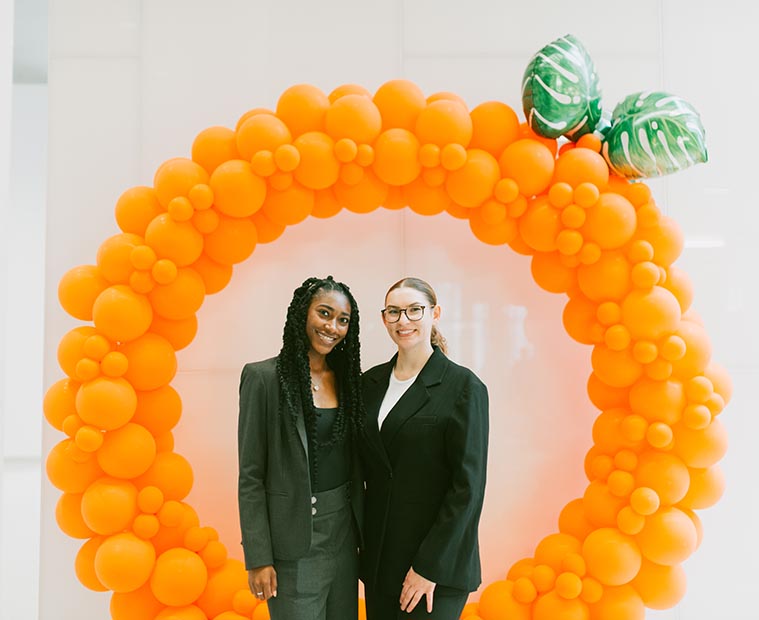 two students stand under a balloon arch that looks like an orange made up of a lot of orange balloons