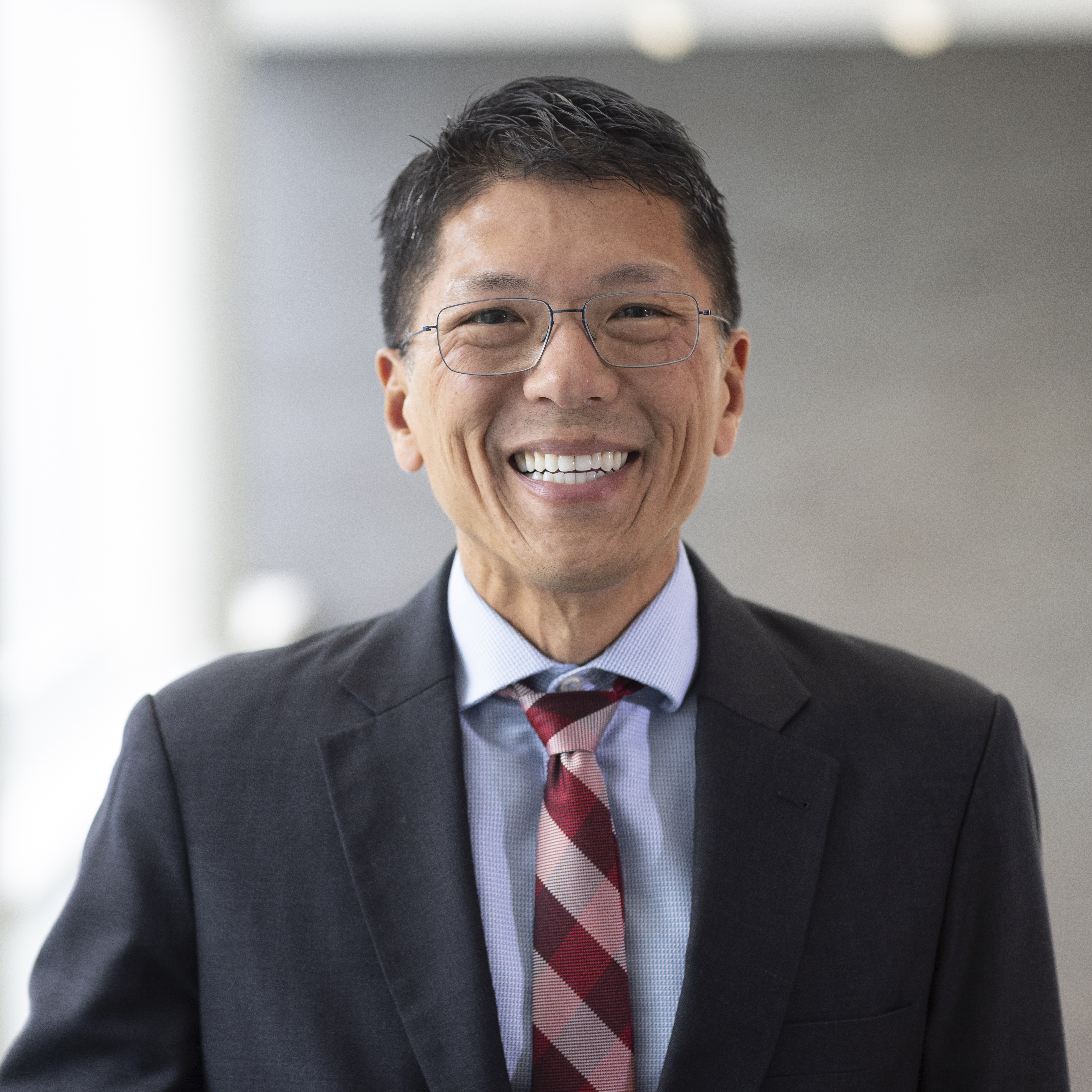 Headshot of Dean Terence Lau wearing a black suit in front of an abstract gray background.