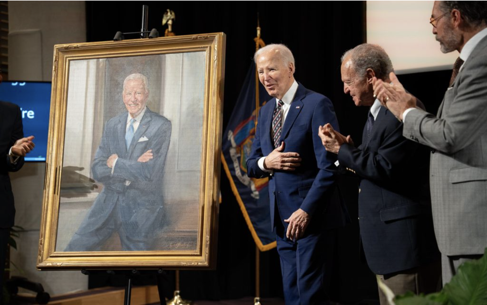 President Joe Biden smiles next to his painted portrait, while three men on stage applaud him.