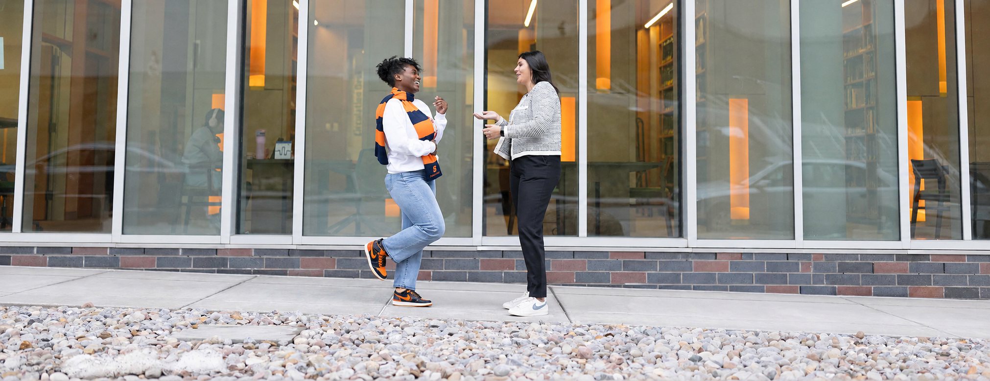 Two students stand in front of the College of Law talking to each other, one wearing a scarf and orange shoes and the other in a gray jacket.