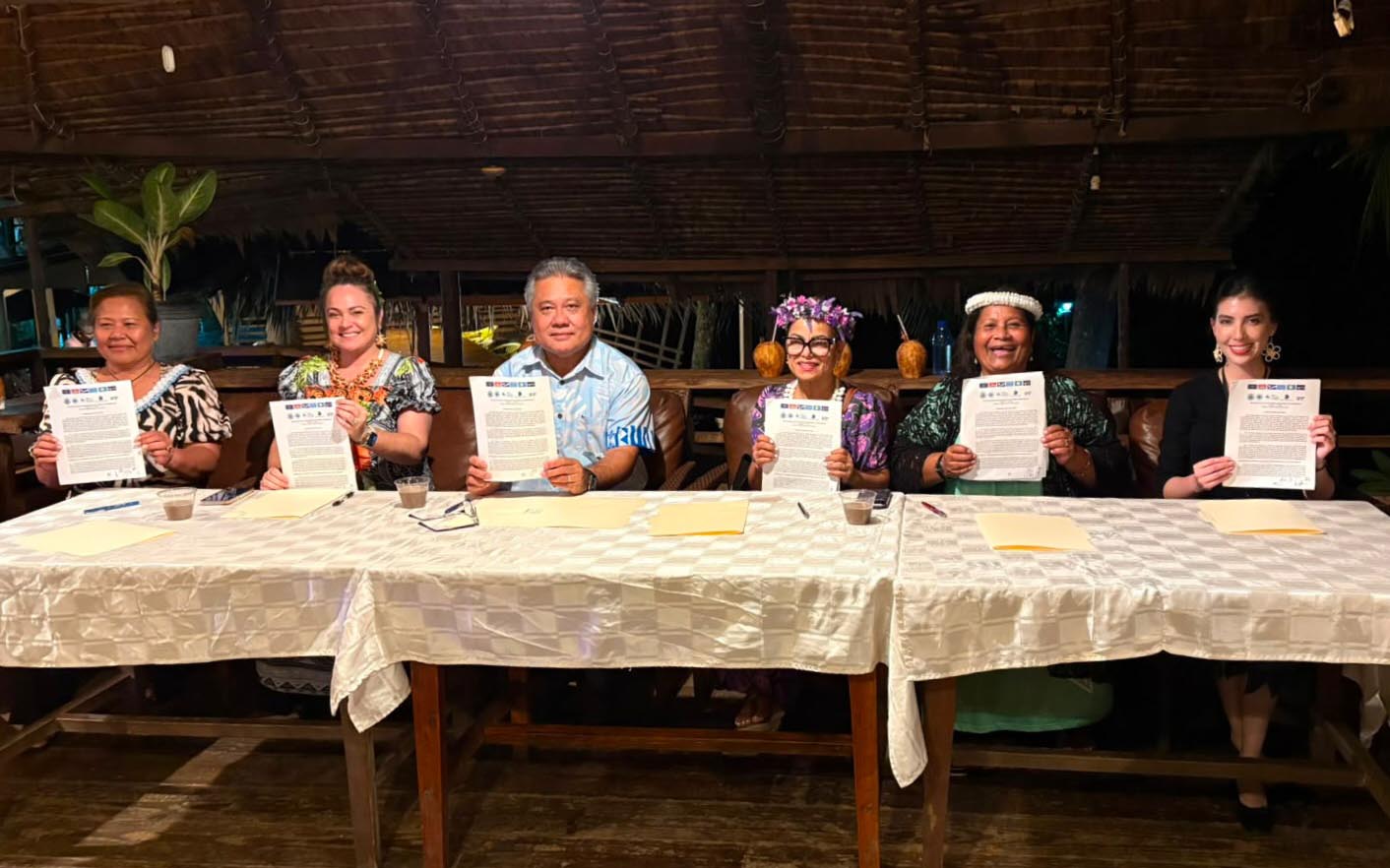 Sarah sits at a table with 5 fellow ministers at the signing of their declaration, holding up their papers.