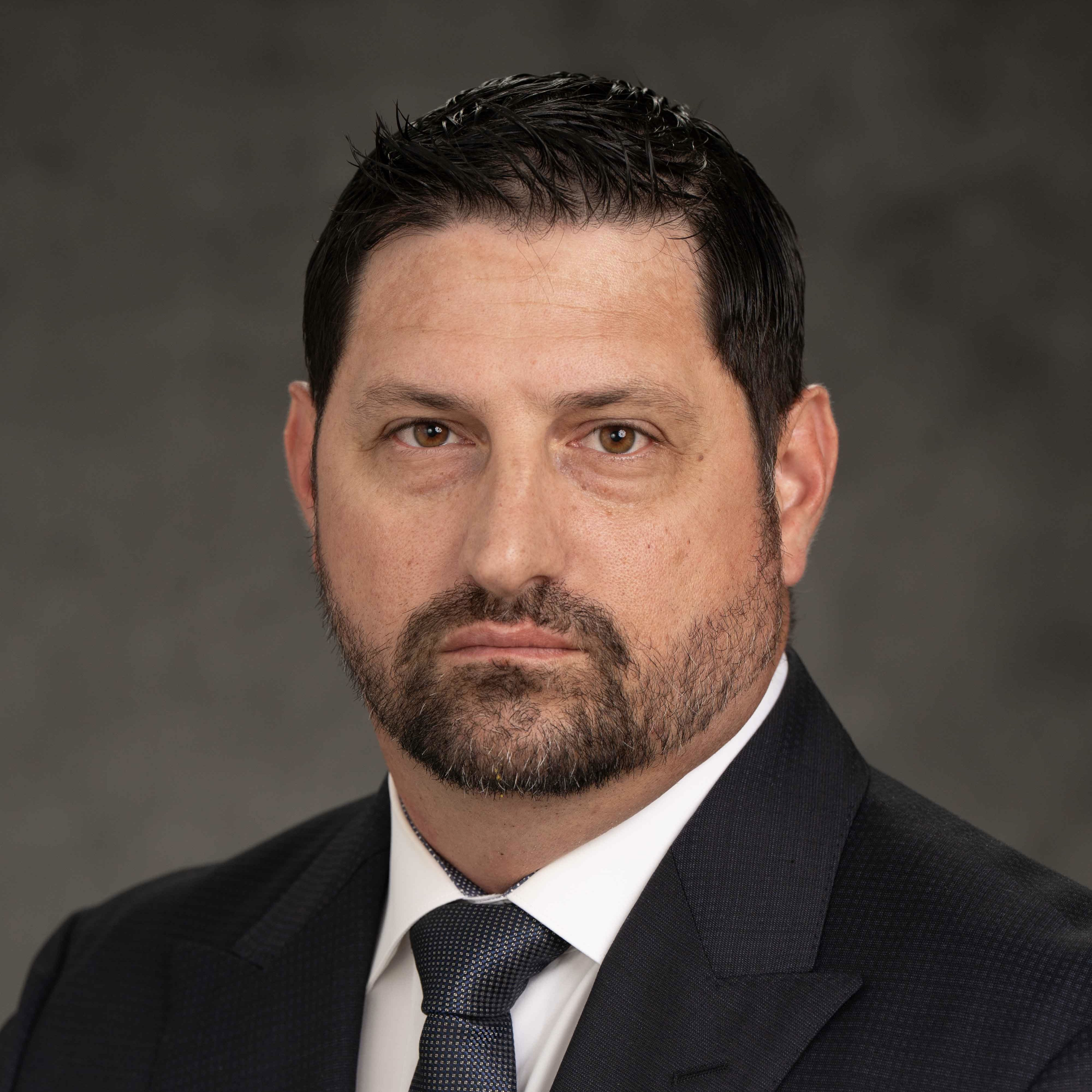 Timothy Riffle wears a black suit and white shirt in front of a gray background.