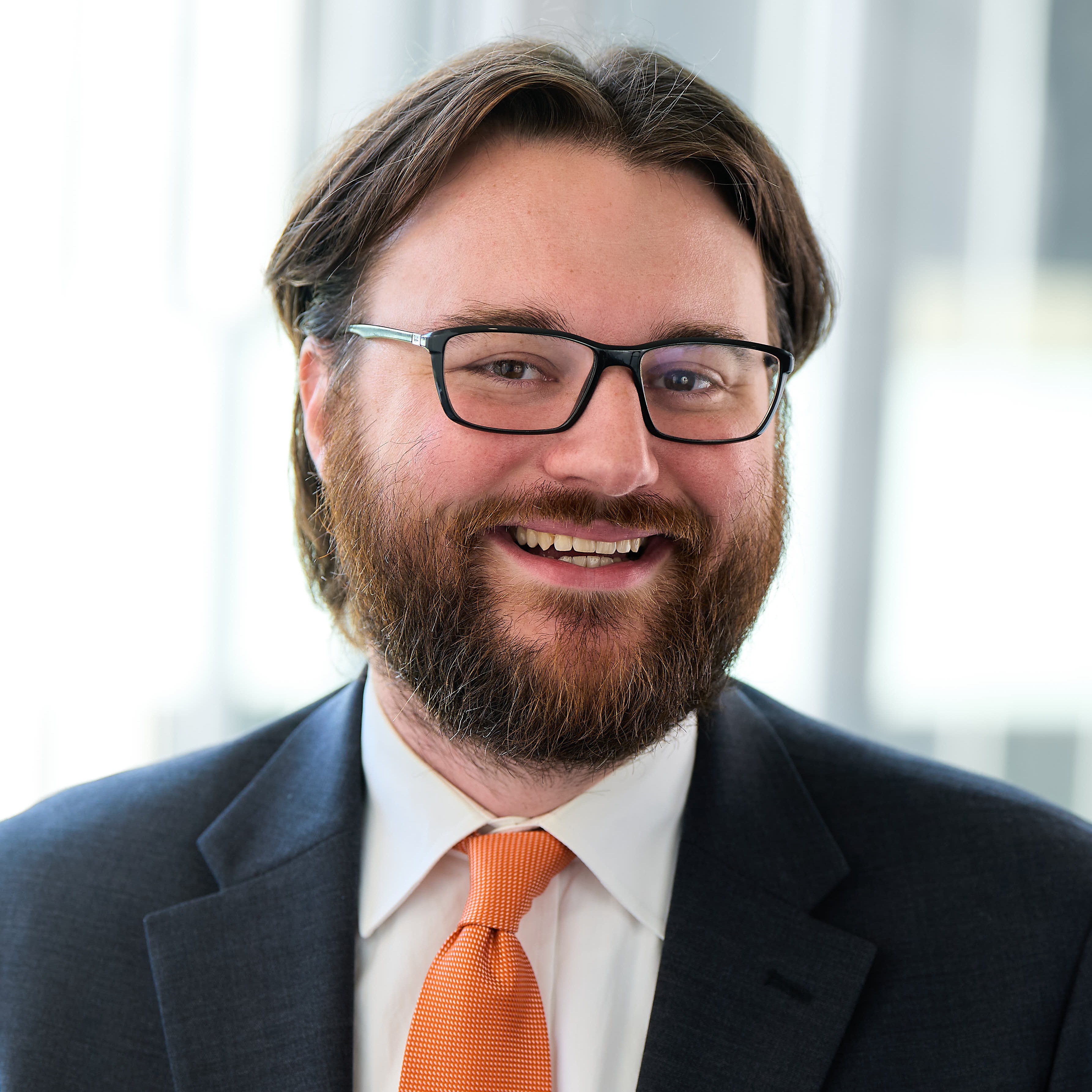 A man with dark hair and glasses wearing a dark suit and orange tie.