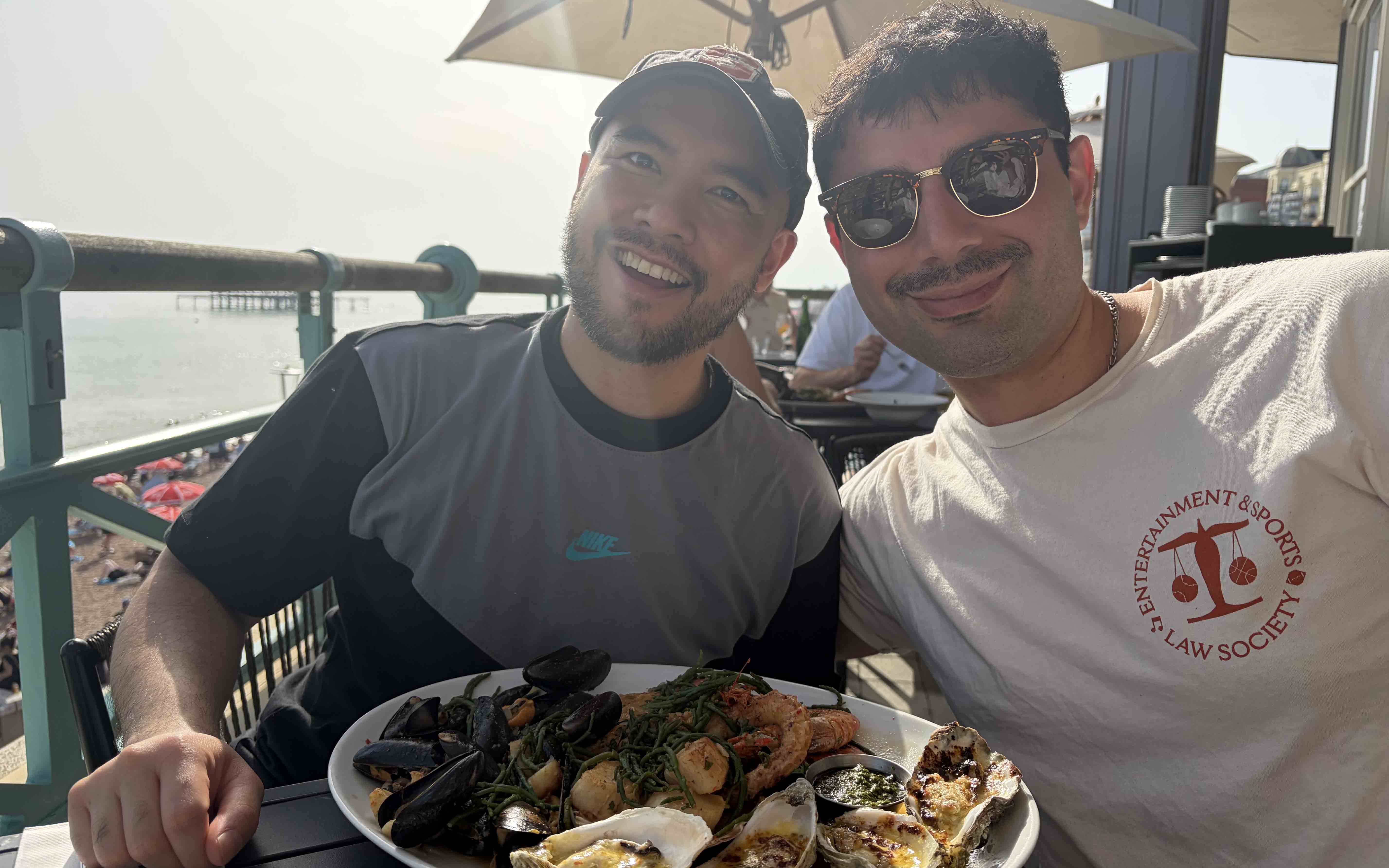 Two students smile in front of a plate of oysters at a restaurant, wearing t-shirts.