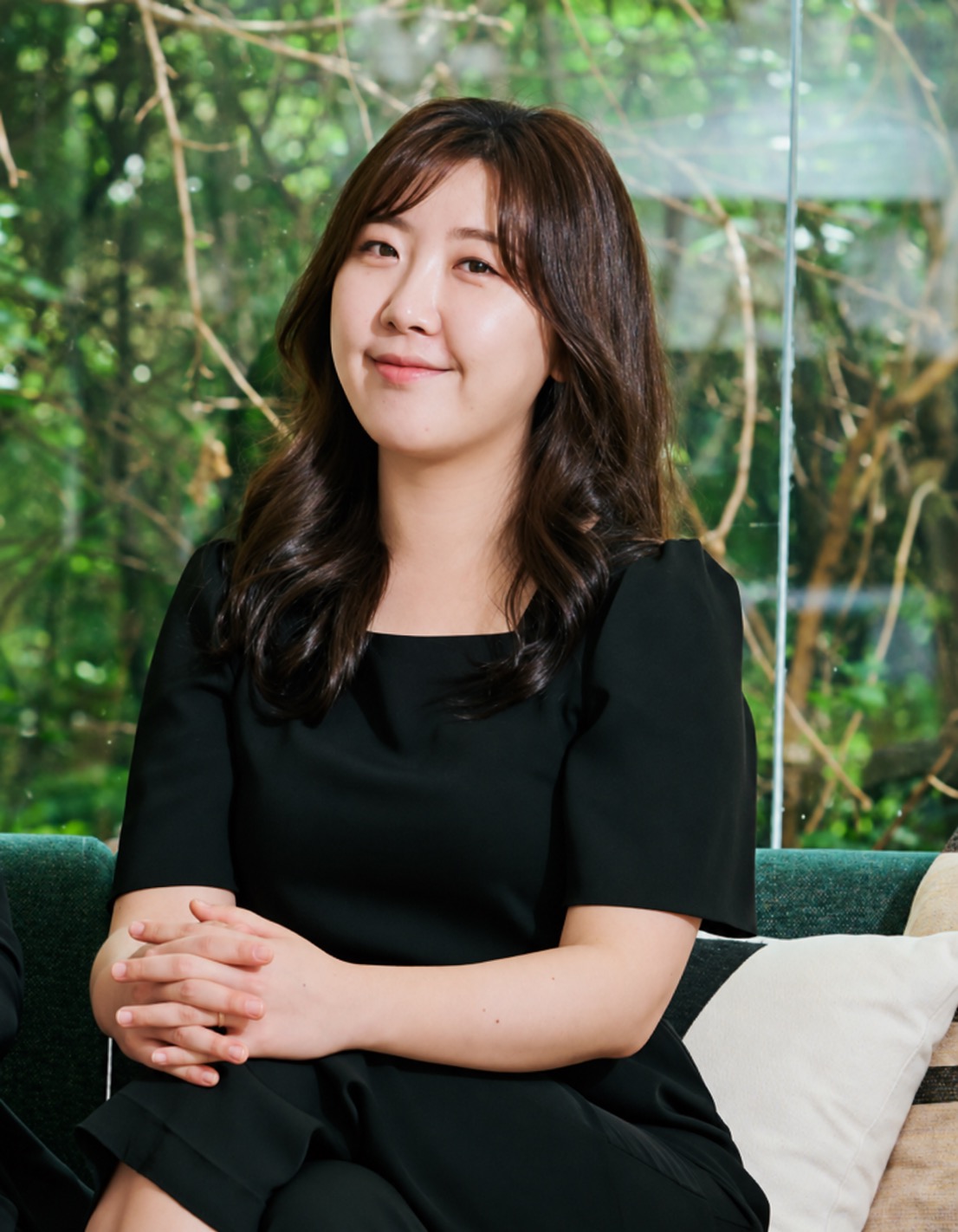 Jenny Oh smiles wearing a black dress in front of some green trees.